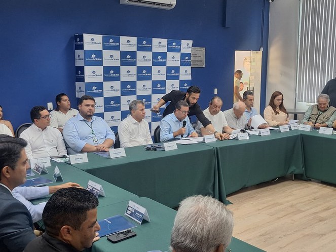 Foto mostra homem falando ao microfone em um evento com outras pessoas