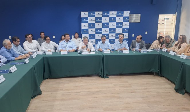 Foto mostra homem falando ao microfone em um evento público com outras pessoas