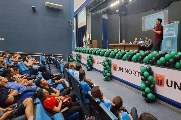 Foto mostra homem de blusa vermelha com microfone na mão e falando a um grupo de acadêmicos