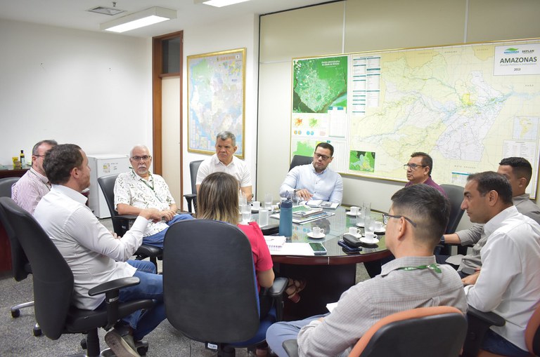 Foto mostra pessoas reunidas em uma mesa redonda, com quadros de mapas ao fundo