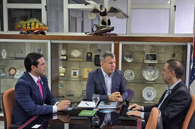 Foto mostra três homens vestidos de paletó, durante reunião com a imagem de um peixe um pássaro de fundo  