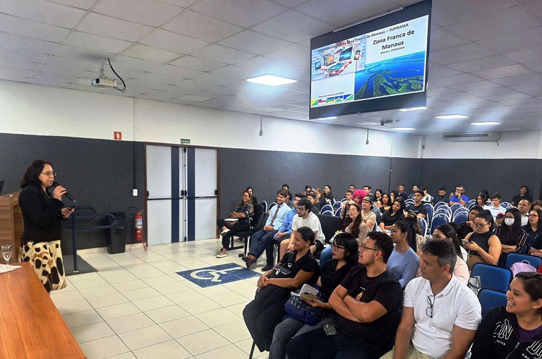 Foto mostra mulher de blazer preto e microfone na mão falando a estudantes universitários