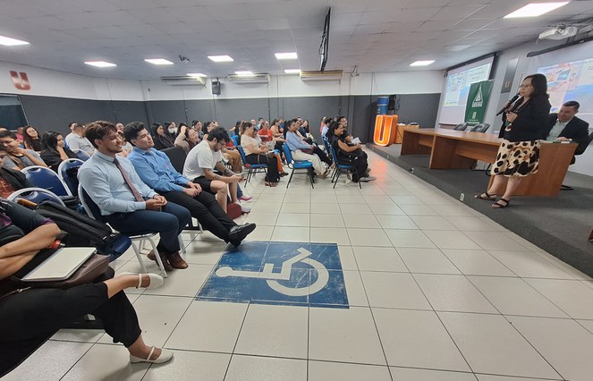 Foto mostra mulher de blazer preto e microfone na mão falando a universitários