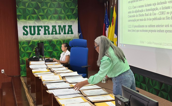 Foto mostra mulher de blusa verde arrumando documentos, com outra mulher ao fundo utilizando o computador