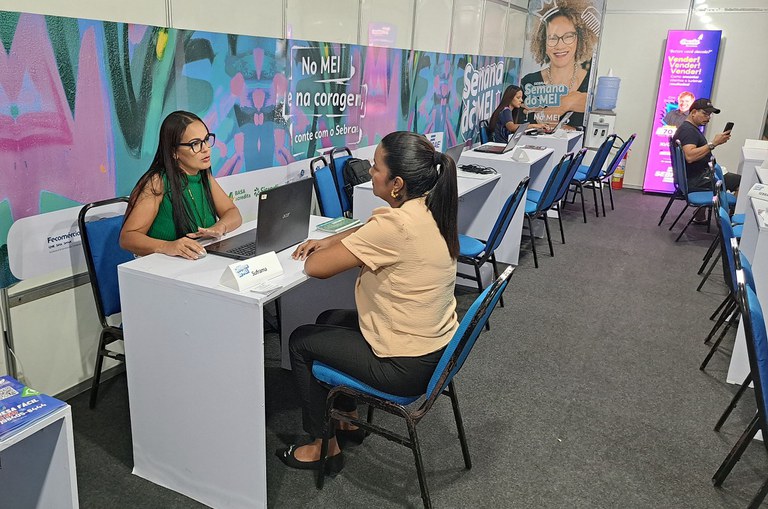 Foto mostra de óculos e blusa verde dando uma explicação a outra mulher