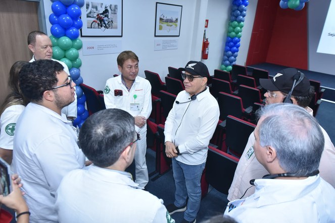 Foto mostra homem de boné preto e óculos escuros conversando com outros homens, num ambiente com balões e um quadro de uma moto ao fundo