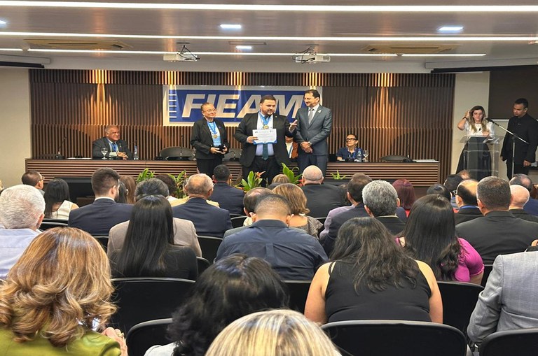 Auditório cheio durante cerimônia na FIEAM. No palco, cinco pessoas em pé, uma delas segura certificado, enquanto outras assistem sentadas. Banner da FIEAM ao fundo.