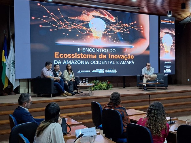 Foto mostra pessoas num evento, com um telão ao fundo