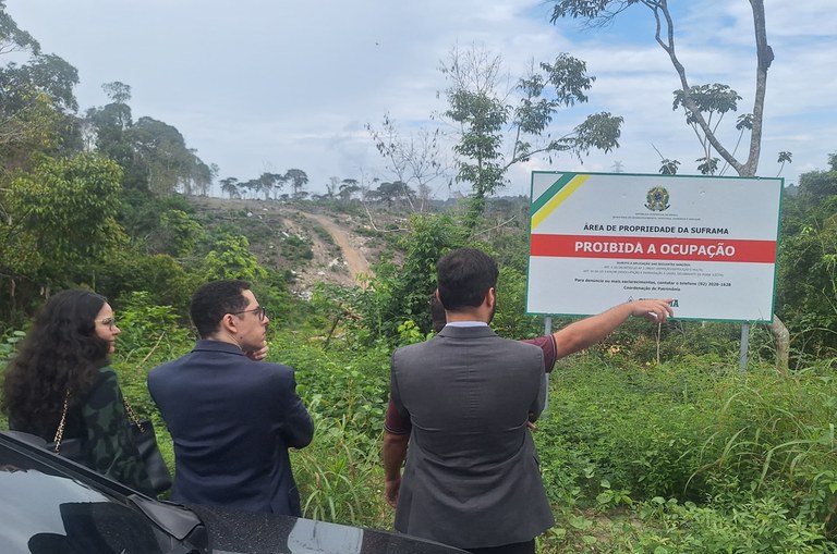 Foto de membros do ministério público e da Suframa inspecionando um terreno no Distrito Agropecuário. Neste terreno há uma placa alertando que a área é terreno da Suframa e é proibida a ocupação.