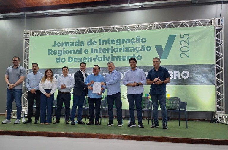 Imagem apresenta um homem assinando um documento num pulpito em frente a um público em um auditório