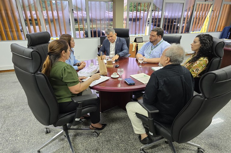 Foto mostra homem de blazer azul mexendo num papel enquanto escuta explicação de uma mulher sentada à direita dele