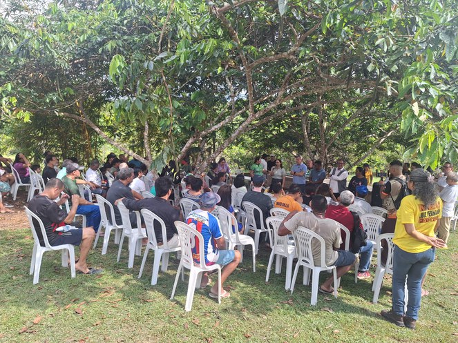 Foto mostra pessoas numa reunião a céu aberto em um terreno e sentadas