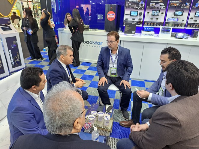 Foto mostra homem de paletó azul fazendo uma explicação a outros homens, sentados com ele em um estande de uma feira de eletroeletrônicos