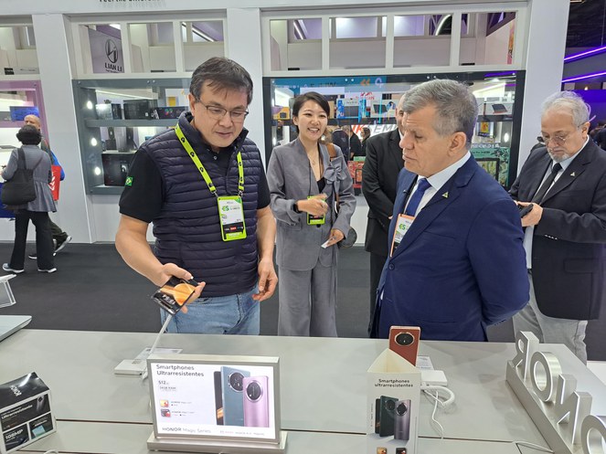 Foto mostra homem de óculos e crachá grande no peito, mostrando um produto a outro homem, ambos observados por uma mulher ao fundo