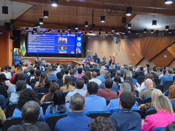 Foto mostra um auditório lotado num evento e um telão ao fundo