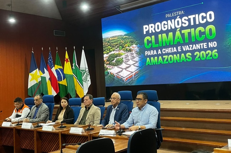 Foto mostra pessoas sentadas num evento com um painel grande ao fundo