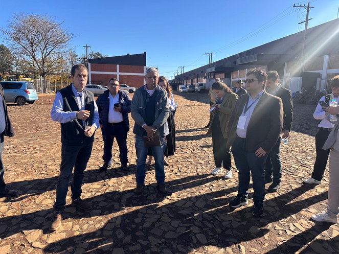 Homem discursa do lado de fora de uma construção, ao sol, enquanto outras pessoas, em volta dele, observam.