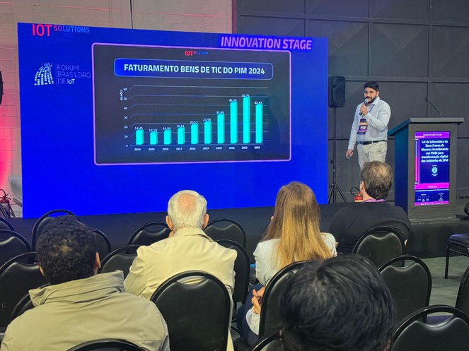Foto mostra homem com microfone na mão falando em público com o apoio de um painel