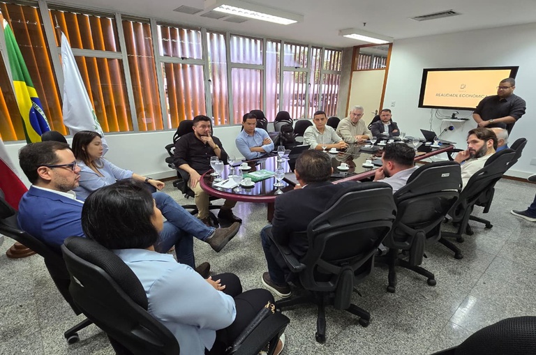 Pessoas em uma sala pequena sentadas em volta de uma mesa oval vendo uma apresentação ao fundo