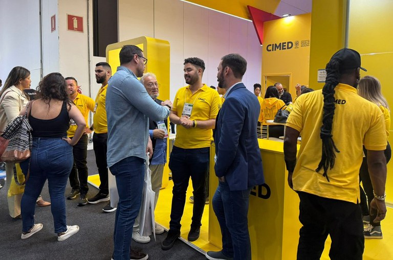 Foto mostra homem de blusa azul clara em conversa com outros homens em um ambiente de fundo amarelo