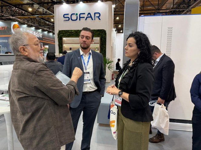 Foto mostra homem de jaqueta marron falando com duas pessoas