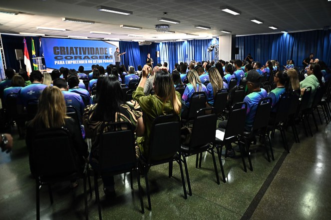 Foto mostra homem em pé falando para um auditório lotado, com um grande banner ao fundo