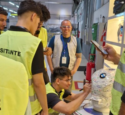 Foto mostra jovem de jaqueta escrevendo num equipamento, observado por outras pessoas