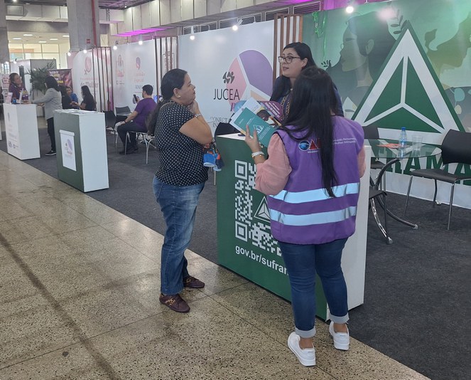 Foto mostra mulher vestindo blusa azul e usando óculos prestando atendimento num estande de um evento