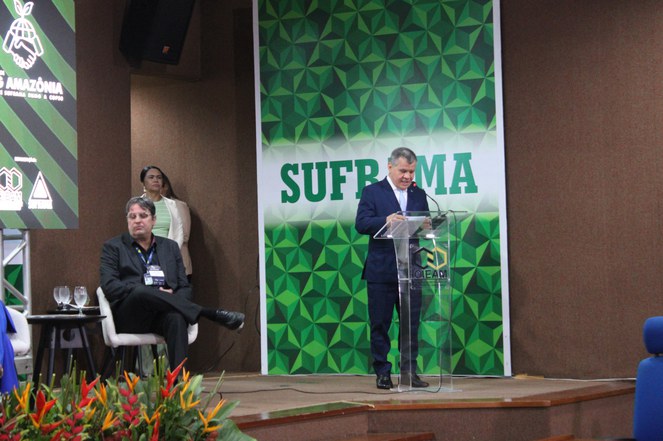 Foto mostra homem de paletó escuro segurando microfone e falando em encontro