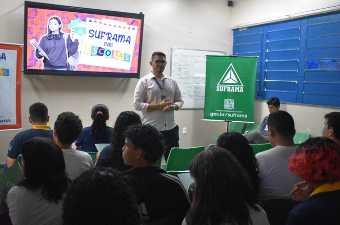 Foto mostra homem de blusa branca falando a um grupo de estudantes com um telão ao fundo