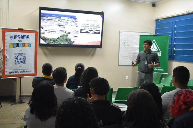 Foto mostra homem de blusa cinza escuro falando a um grupo de estudantes com um telão ao fundo