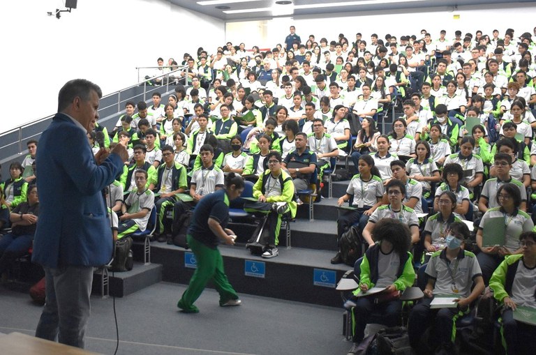 Foto mostra homem de paletó e calça azul falando para um auditório cheio de estudantes