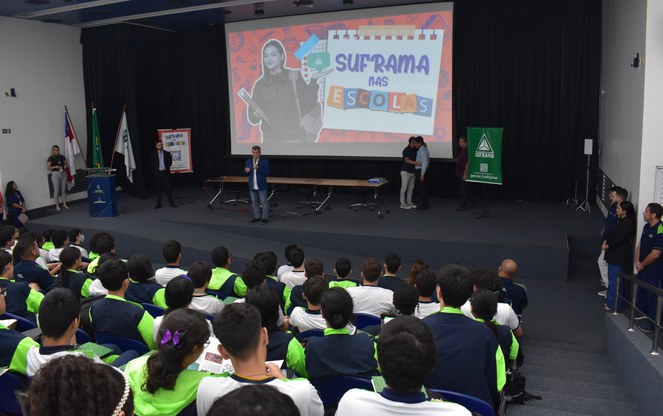 Foto mostra um homem de paletó e calça azul falando para um auditório cheio de estudantes