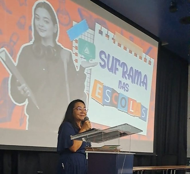 Foto mostra mulher falando com microfone na mão e um grande painel de fundo com os dizeres Suframa nas Escolas