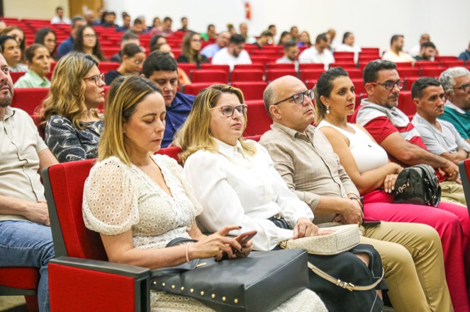 Foto mostra mulher olhando para o celular, enquanto outras pessoas assistem atentas a uma apresentação