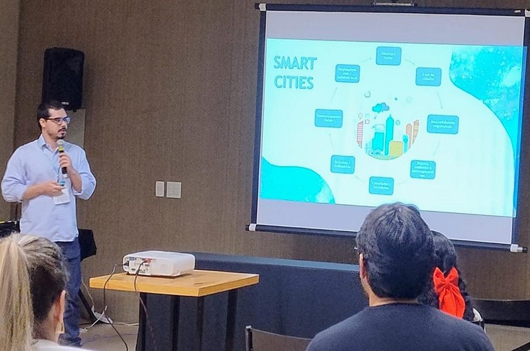 Foto mostra homem de óculos e microfone na mão ministrando uma palestra com um telão de apoio