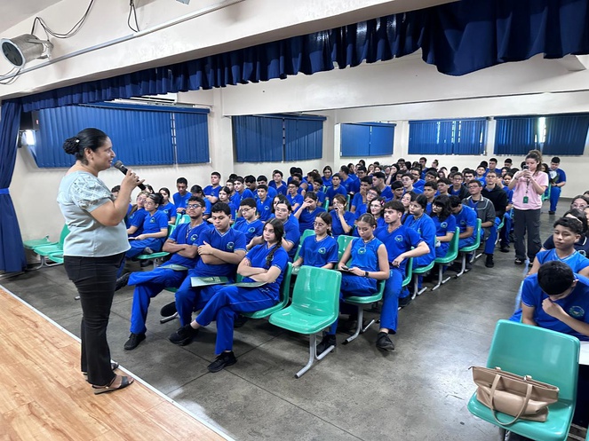 Foto mostra mulher em pé fazendo uma apresentação a um grupo de alunos