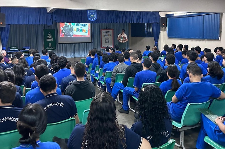 Foto mostra mulher em pé explanando para uma sala cheia de alunos e com um painel grande como apoio