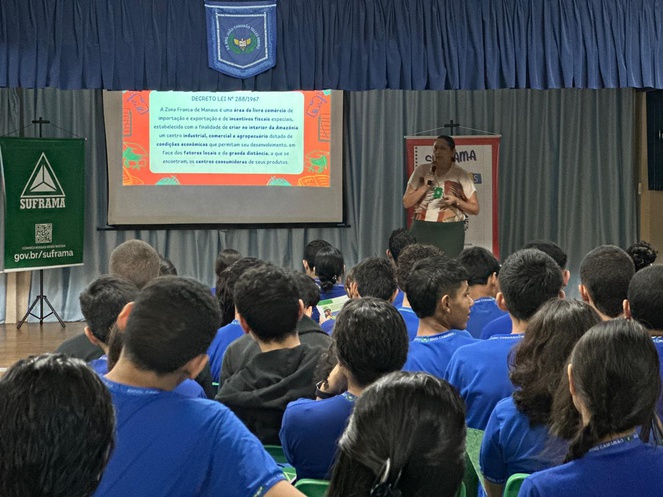 Foto mostra uma mulher em pé explanando para uma sala cheia de alunos e com um painel grande como apoio