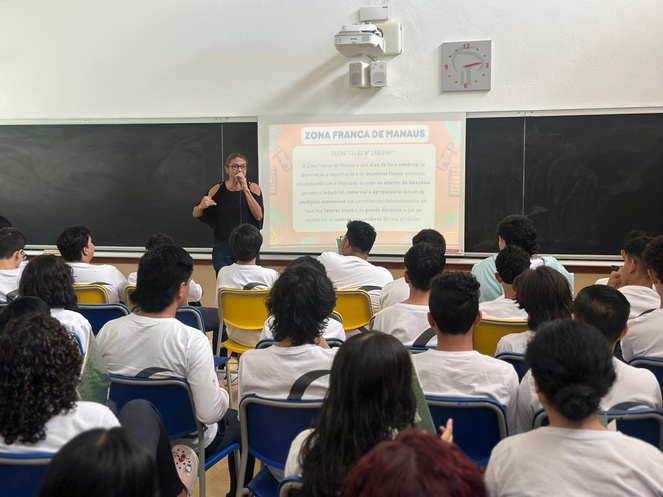 Foto mostra mulher ministrando uma palestra a estudantes com o apoio de um telão