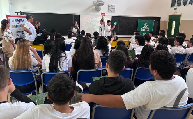 Foto mostra mulher apresentando uma dinâmica após palestra a estudantes com o apoio de um telão