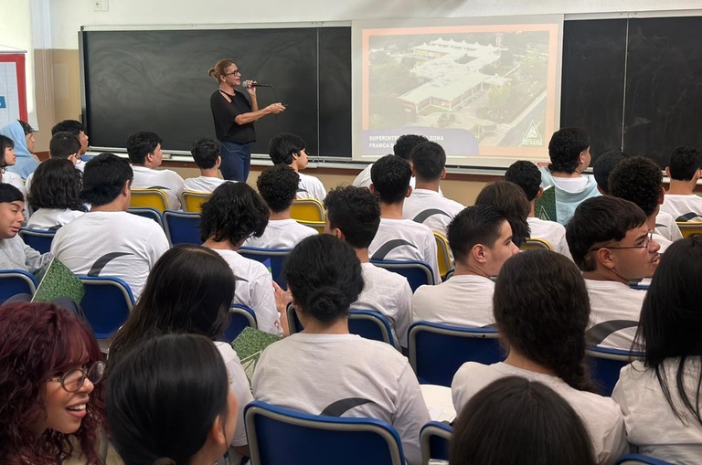 Foto mostra uma mulher de óculos, em pé e com microfone na mão apontando para um telão, com estudantes como espectadores