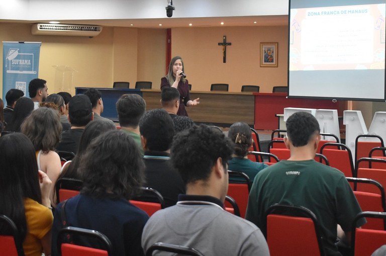 Foto mostra mulher falando ao microfone a grupo de universitários, com um telão como apoio