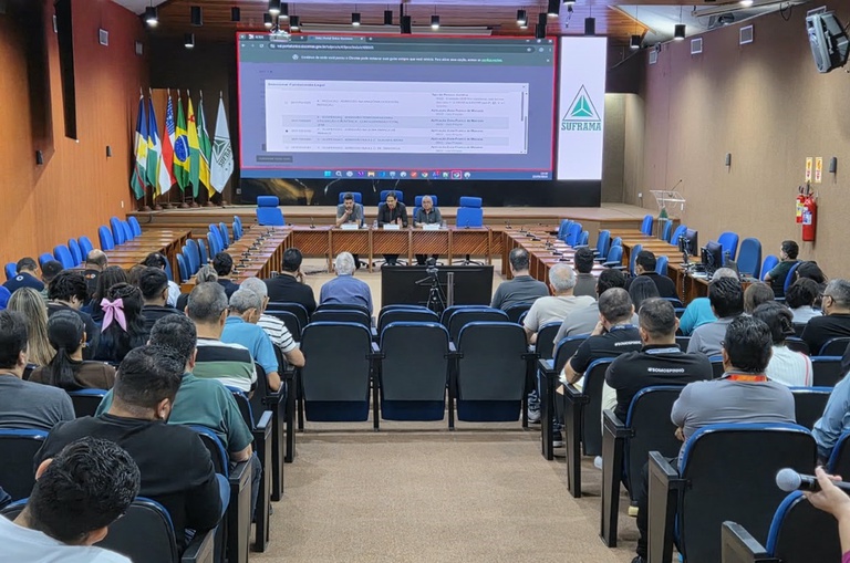 Foto mostra três pessoas numa bancada durante palestra, observadas por outras e tendo um telão como apoio