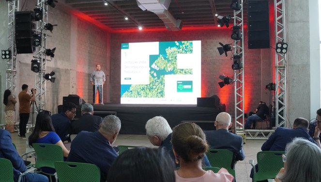 Foto mostra homem de blusa marrom falando em evento público