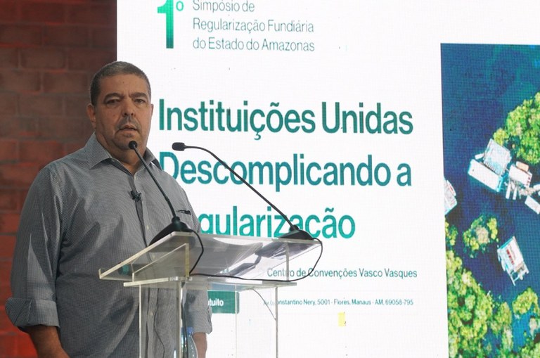 Foto mostra homem de blusa marrom falando ao microfone