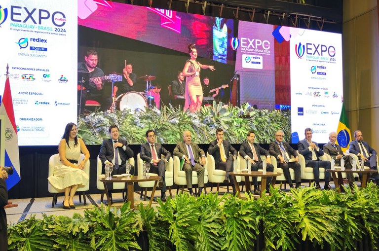 Foto mostra grupo de pessoas sentadas num grande evento e com um painel ao fundo