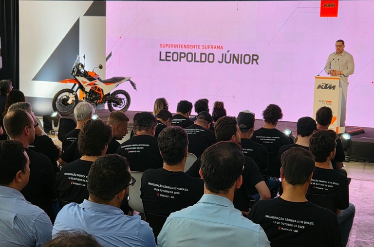 Foto mostra homem falando num evento público, com um telão como apoio