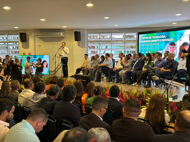 Foto mostra homem falando em evento público, com painéis eletrônicos ao fundo