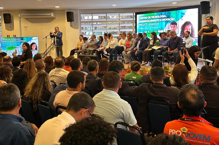 Foto mostra homem falando em público, durante evento concorrido, com pessoas sentadas acompanhando e paineis eletrônicos ao fundo